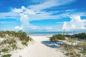 Ein sandiger Küstenpfad führt zum Meer, begleitet von spärlicher Vegetation an beiden Seiten.