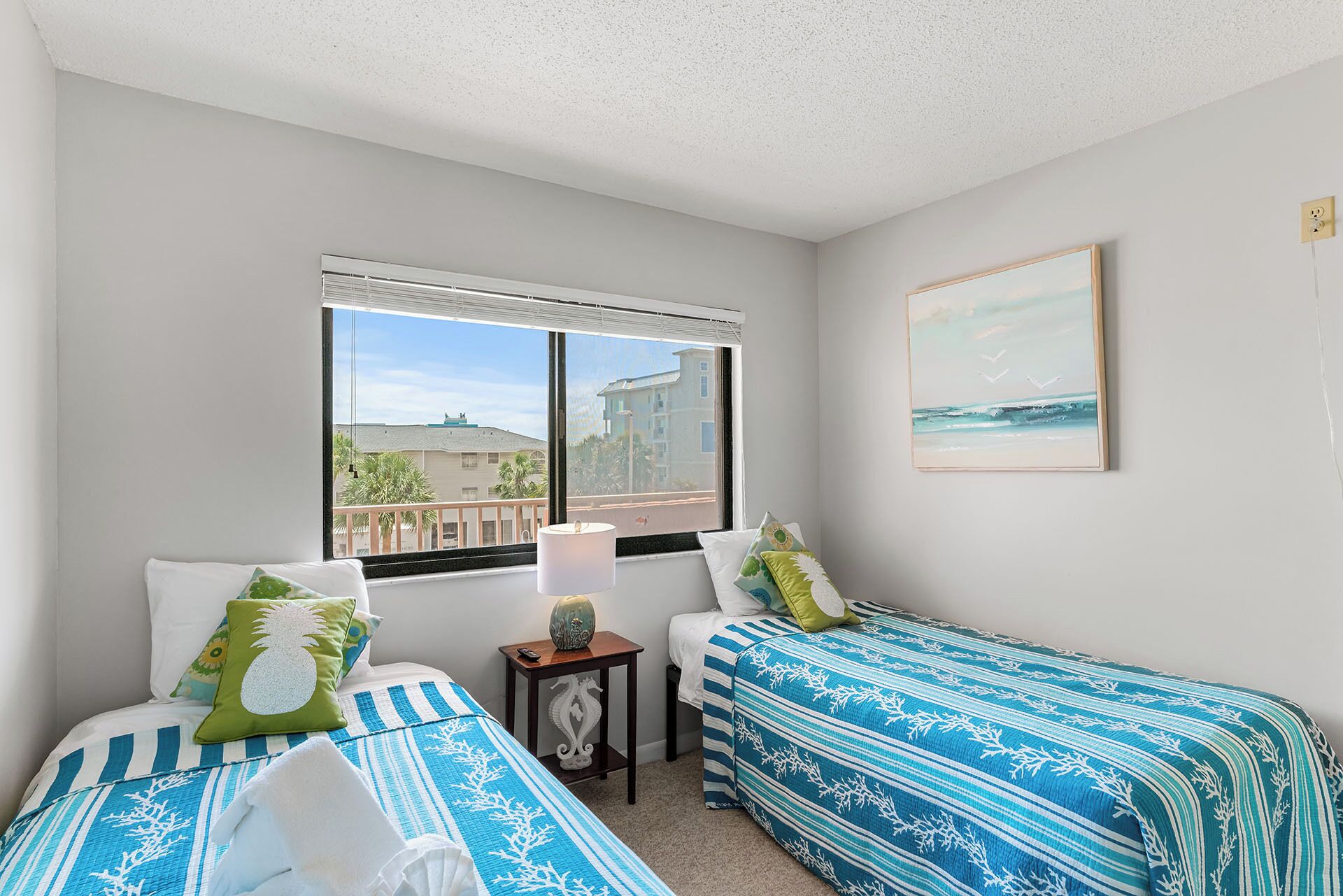 A bedroom with two beds, a window with a view of buildings, a painting on the wall, and a small table with a lamp.