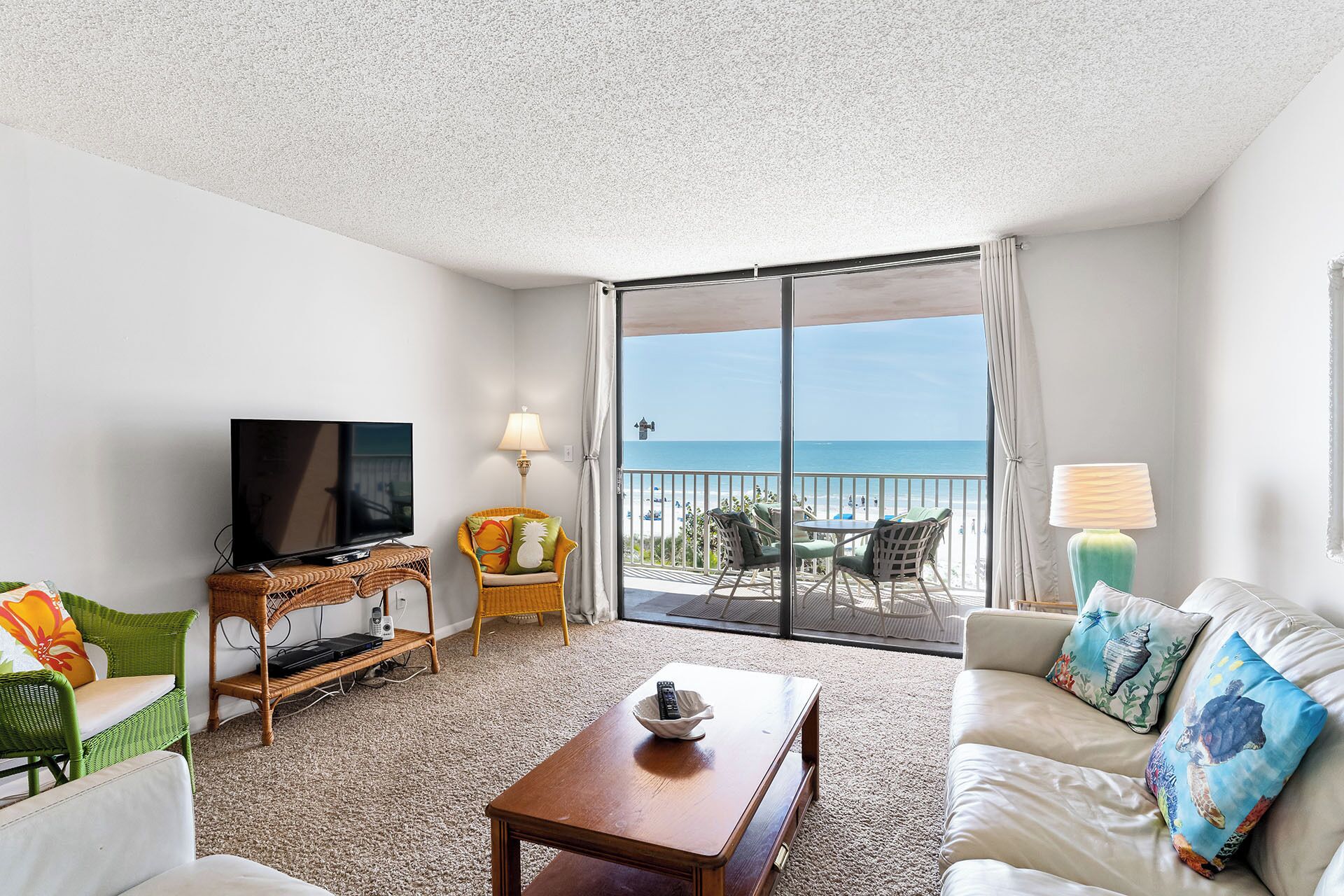 A hotel room with a TV, a wicker side table, a sofa with decorative pillows, a wooden coffee table, and a balcony with a view of the beach.