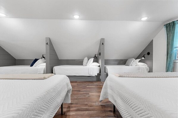 A modern, well-lit room with three beds arranged in a U-shape, wooden flooring, and a sloped ceiling.