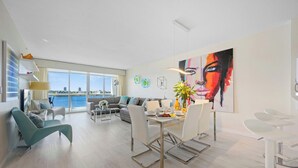 A modern living room with a sofa, dining table, and chairs. There is a large window with a view of the water. - Comfortable condo with amazing bay views | WIFI + Parking (Sunny Isles Beach)