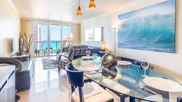 A modern dining area with a glass table, chairs, and a view of the ocean through large windows. - Warm condo w/ striking ocean & park views | WIFI + Parking (Sunny Isles Beach.)