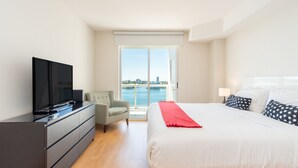 Moderni hotellihuone, jossa on suuri sänky, televisio lipaston päällä, parveke, jolta on näkymä vesistöön, ja kaupunkimaisema kauempana.