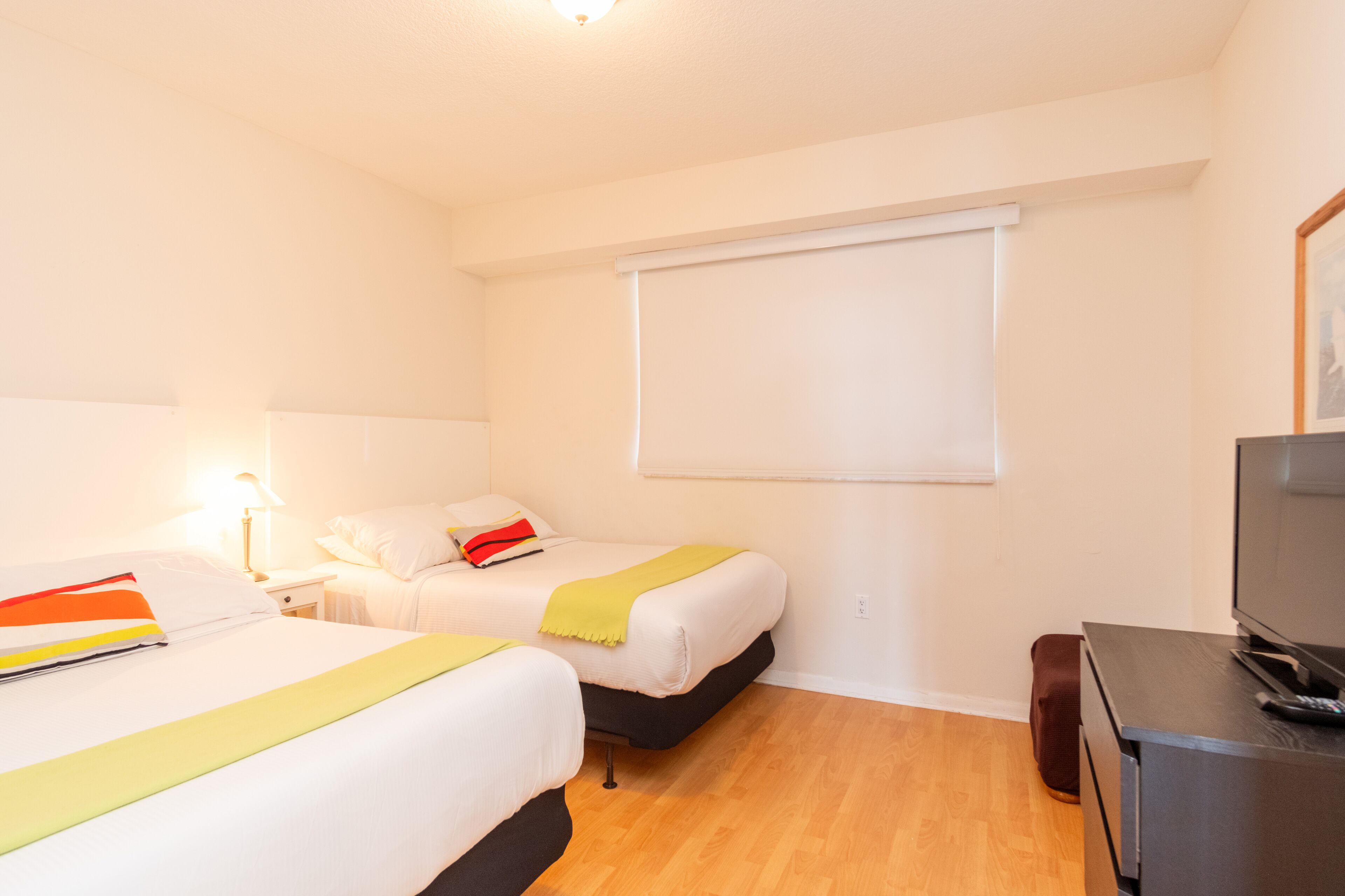 A hotel room with two beds, a television, a desk, and a window with blinds.