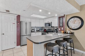 A modern kitchen with white cabinets, stainless steel appliances, a large island with bar stools, and a wall clock.