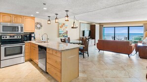 A modern kitchen with stainless steel appliances, a dining area with a view, and a living room with a sofa and a balcony overlooking the ocean.