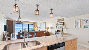 A kitchen with granite countertops, stainless steel sink, and a dishwasher. There are pendant lights, a view of the ocean, and a 'Hello Sunshine' sign on the wall.
