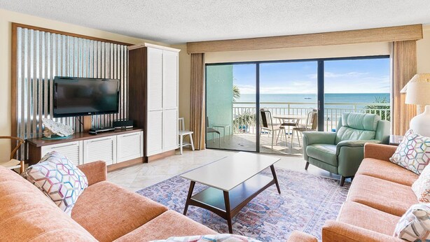 A spacious living room with a flat-screen TV, a sofa set, a coffee table, and a balcony with a view of the ocean.