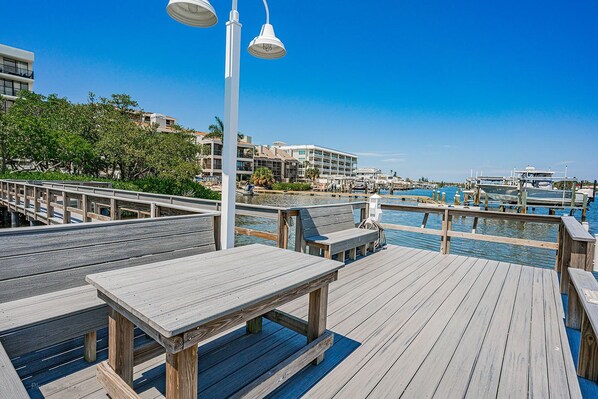 A wooden deck with benches and tables, overlooking a marina with boats and a clear blue sky.