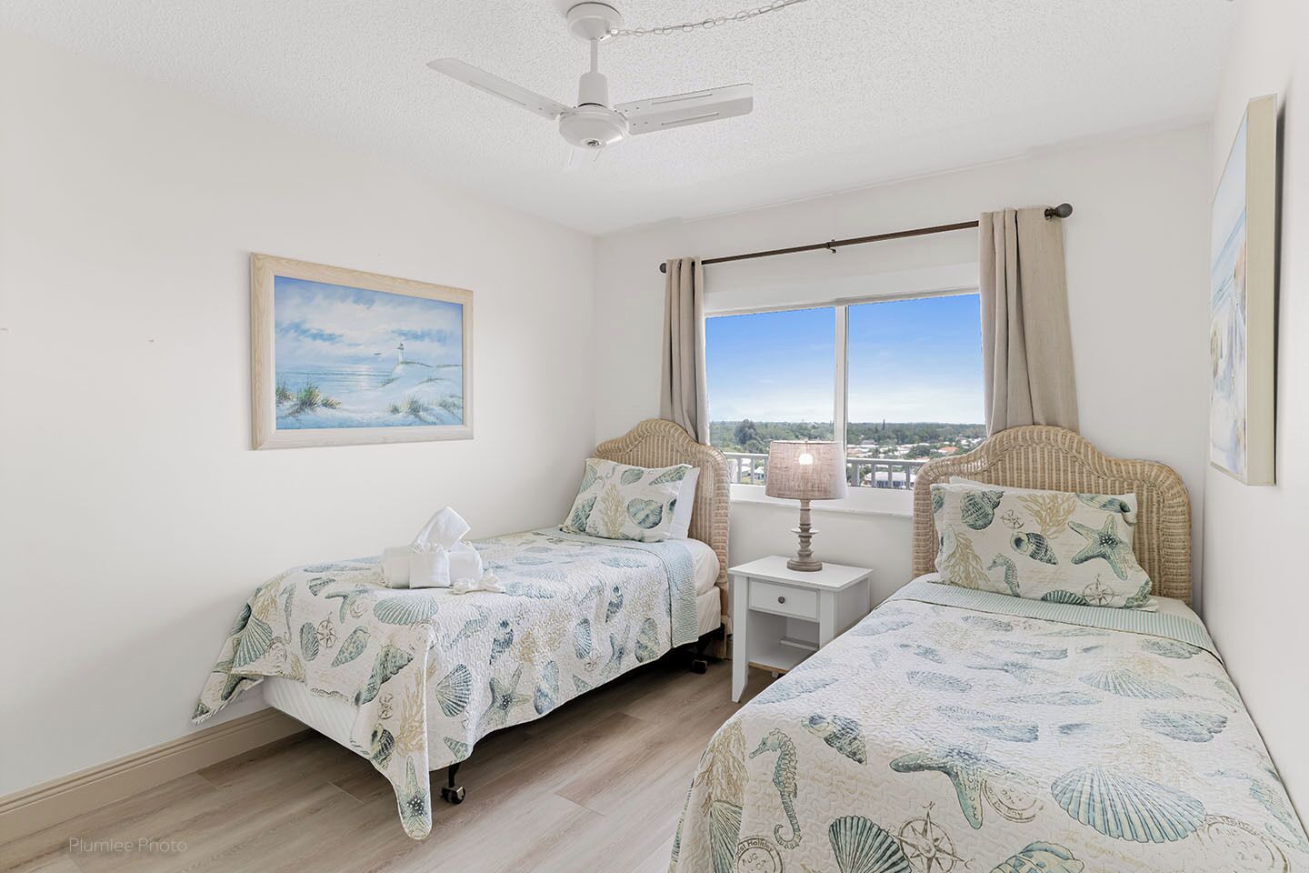 A room with two beds, a ceiling fan, a window with a view, and a painting on the wall.