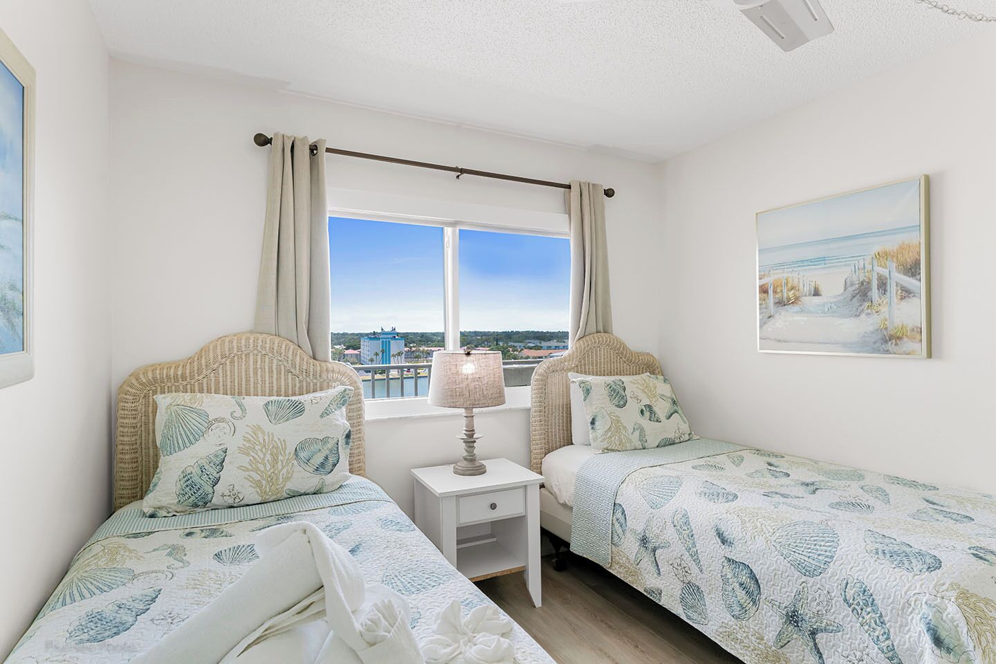A bedroom with two single beds, a nightstand, a lamp, a window with curtains, and a beach-themed painting.