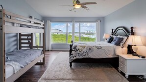 Une chambre avec un lit superposé, un lit simple, un ventilateur de plafond, une table de chevet avec une lampe, et une vue sur la verdure à l’extérieur.