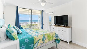 A bedroom with a bed, a TV, a ceiling fan, and a view of the sea.