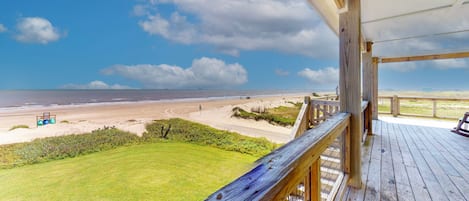 A beach view from a wooden deck with a grassy lawn and a clear sky.