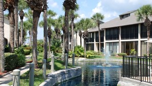 A pool area with palm trees, a fountain, and a building with large windows. - Soak up the Florida sunshine in this modern Gold-rated condo overlooking the lagoon and pool. (Saint Augustine)