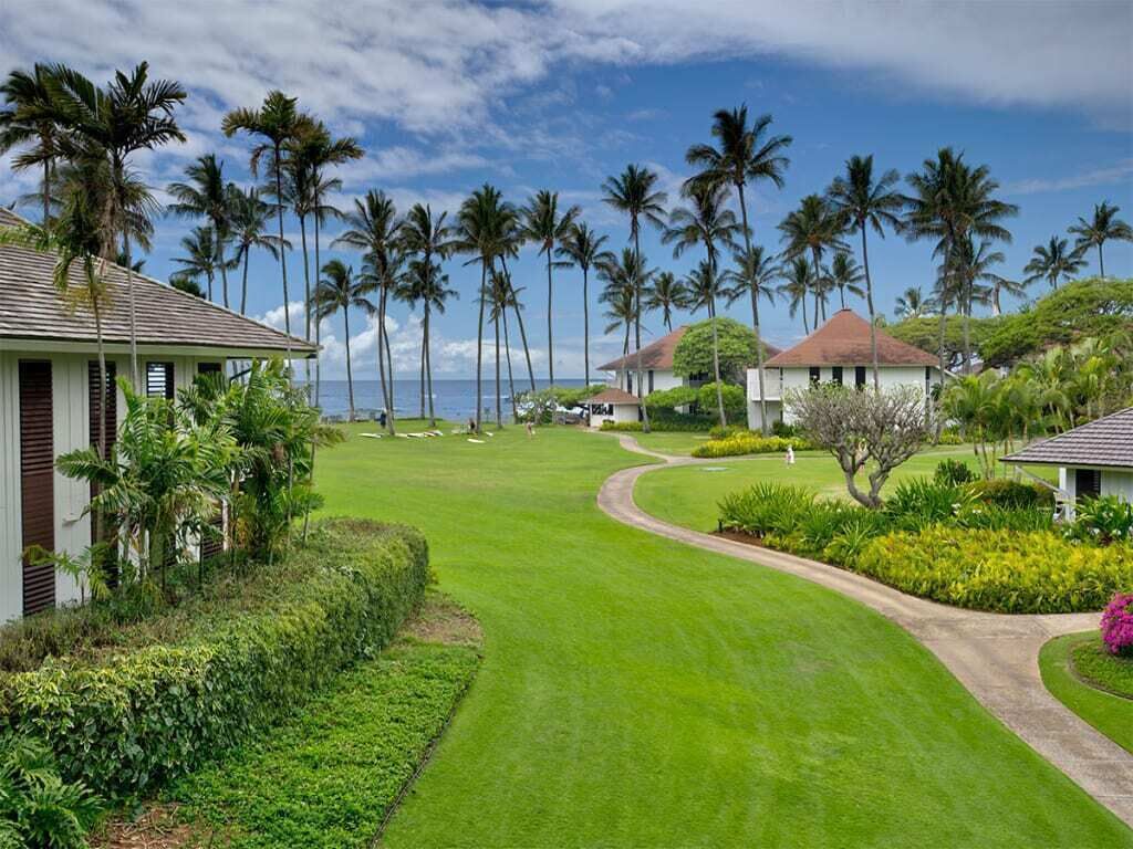 A golf course with a clear path, surrounded by lush greenery and palm trees, leading to a building with a red roof.