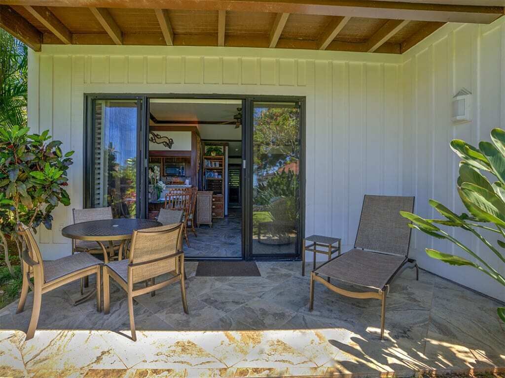 An outdoor patio with a round table and chairs, a lounge chair, and a sliding glass door leading to an interior space with a bookshelf and a television.