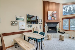 A cozy room with a wooden bench, a table, and chairs. There is a fireplace with a TV above it, and various framed pictures on the wall.
