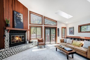 A spacious living room with a fireplace, large windows, and a high ceiling.