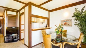 A compact living space with a dining area featuring a glass table and wicker chairs, a television in the corner, and a poster on the wall. - Comforts of Home+Lanai to Lawn, WiFi, Flat Screen, Kitchen–Kiahuna Plantation #2065 (KOLOA)