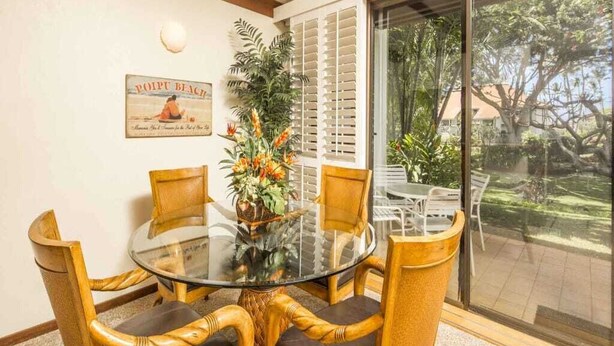 A dining area with a glass table surrounded by wicker chairs with leather cushions, a potted plant, and a poster on the wall. - Comforts of Home+Lanai to Lawn, WiFi, Flat Screen, Kitchen–Kiahuna Plantation #2065 (KOLOA)