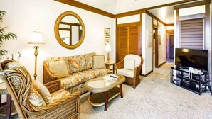 A living room with a sofa, armchair, and coffee table, a television, and a mirror on the wall. - Comforts of Home+Lanai to Lawn, WiFi, Flat Screen, Kitchen–Kiahuna Plantation #2065 (KOLOA)