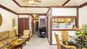 A living room with a sofa, chairs, a glass coffee table, a ceiling fan, and a kitchen area with a microwave. - Comforts of Home+Lanai to Lawn, WiFi, Flat Screen, Kitchen–Kiahuna Plantation #2065 (KOLOA)