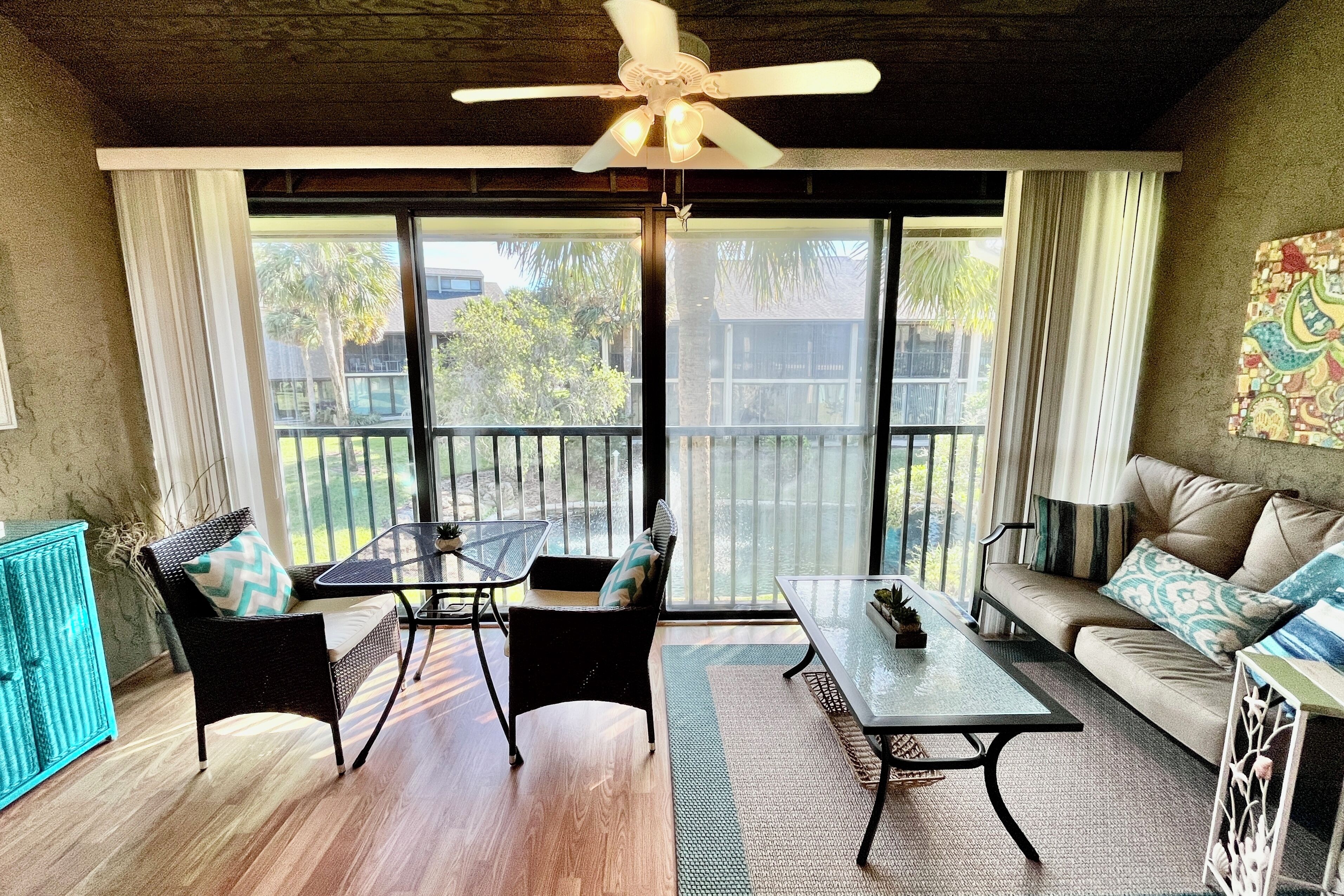 A room with a ceiling fan, a sofa, a dining table, and a chair. There is a view through large windows to the outside.