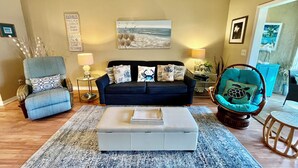 A living room with a dark blue sofa, a light blue armchair, a white ottoman, and a wooden side table with a lamp. - Cozy Silver-rated condo, perfect for enjoying beautiful Florida. (Saint Augustine)