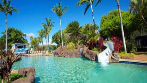 Piscina cristalina con palmeras y una cascada, rodeada de rocas y follaje.