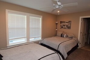 Quarto com duas camas, um ventilador de teto, uma janela com persianas, uma luminária na parede e uma porta.