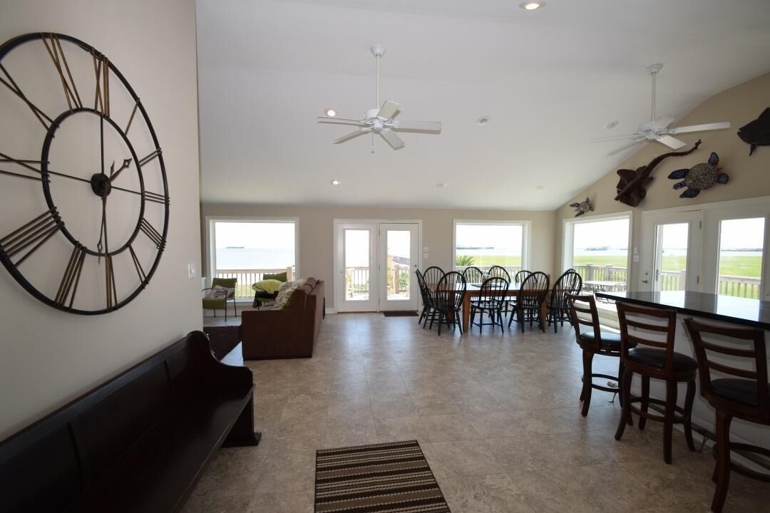A spacious living area with a large wall clock, ceiling fans, a dining area with chairs, and a view through large windows.