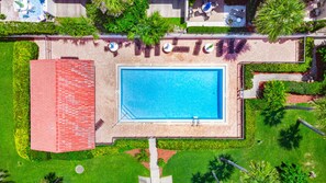 Ein Luftbild eines Schwimmbeckens, umgeben von einem Backsteinhaus, mit einem 'No Diving'-Schild, und einem gepflegten Garten mit verschiedenen Pflanzen und einem Weg zum Pool.