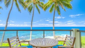 A wooden table and two chairs on a balcony with a view of the ocean and palm trees.