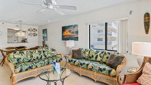A living room with two wicker sofas, a glass coffee table, a ceiling fan, and a large window with a view of a building. - Sand Castle III 201 - GULFFRONT SPIRAL STAIRCASE! (Indian Shores)