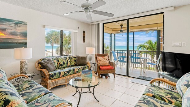 A spacious living room with a large sofa, a coffee table, and a television. There is a balcony with a view of the beach. - Sand Castle III 201 - GULFFRONT SPIRAL STAIRCASE! (Indian Shores)