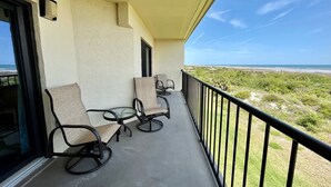 Un balcon avec deux chaises et une table, donnant sur une plage bordée de dunes et sur l’océan.
