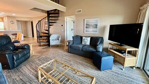 Un salon spacieux avec un canapé en cuir noir, un fauteuil bleu, un grand téléviseur à écran plat et une table basse en bois.