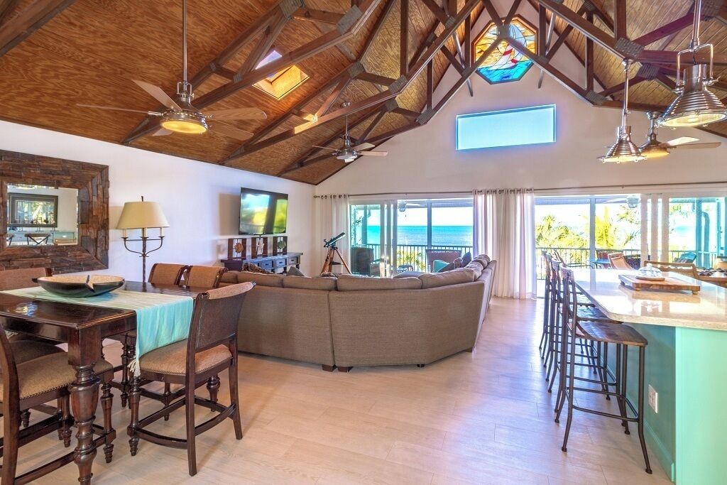 A spacious living area with a large sectional sofa, wooden dining table, and bar stools. There is a large window with a view of the ocean.