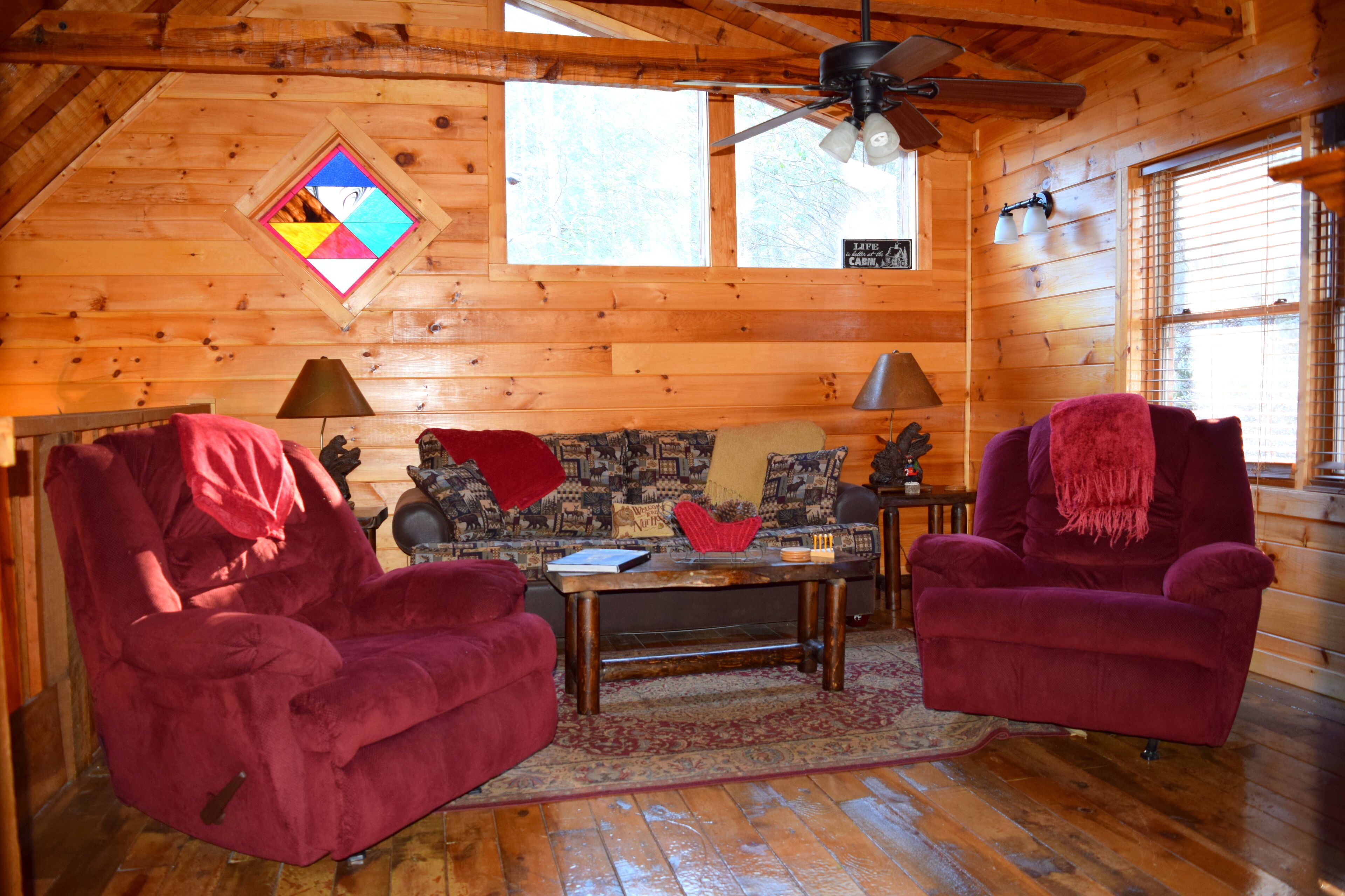 A cozy cabin interior with wooden walls, a ceiling fan, a window with blinds, a sofa, two armchairs, and a small table.