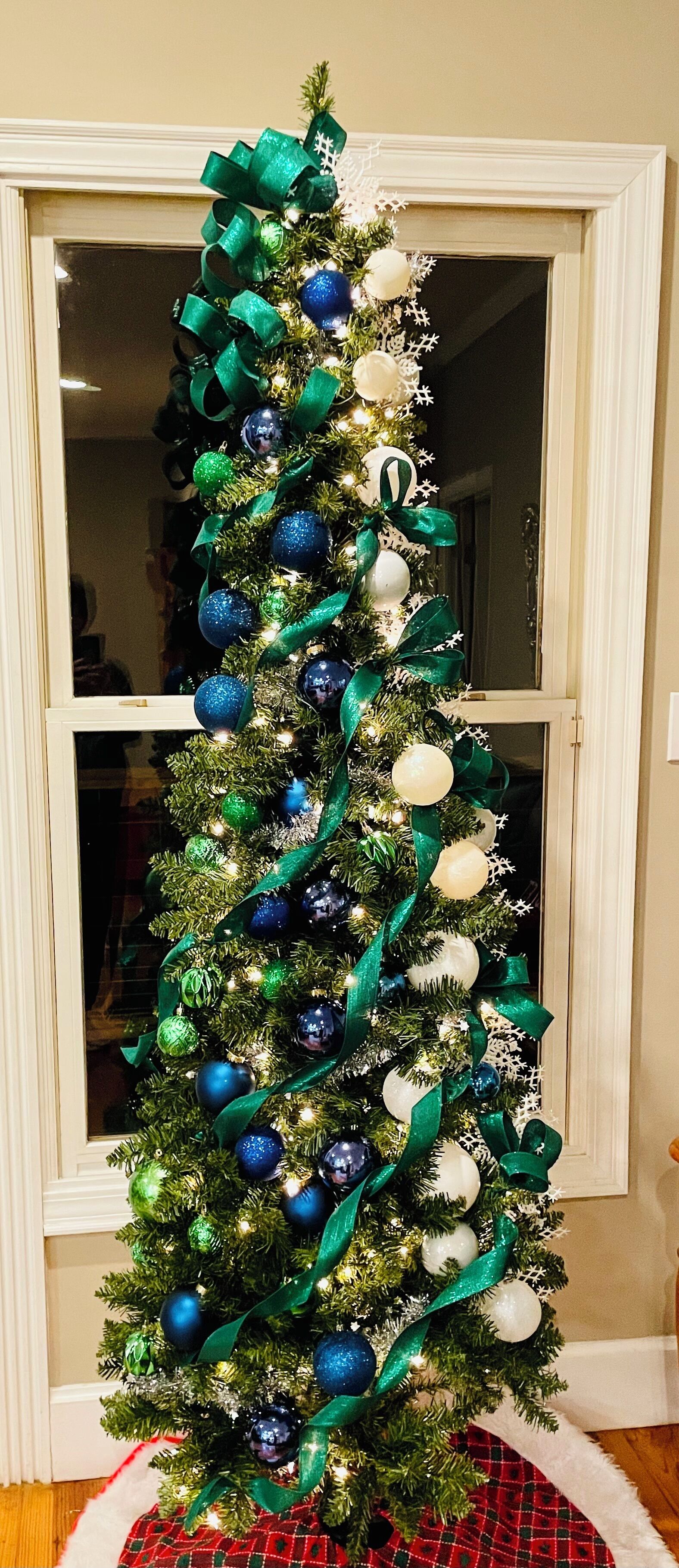 A Christmas tree decorated with blue and white ornaments and green ribbons, placed in front of a window.