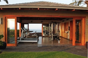 Ein überdachter Outdoor-Fitnessbereich mit Trainingsgeräten, Blick auf das Meer und einer Terrassenfläche.