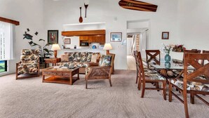A spacious living room with a high ceiling, a dining area with wooden furniture, and a kitchen in the background. - Kona-Keauhou Sunsets+Style! Full Kitchen, Washer/Dryer, WiFi, Lanai, DVD, TV Kanaloa 2106 (KAILUA-KONA)
