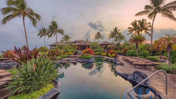 Piscina de un resort rodeada de palmeras y plantas tropicales, con sillas de descanso y un pabellón al fondo.