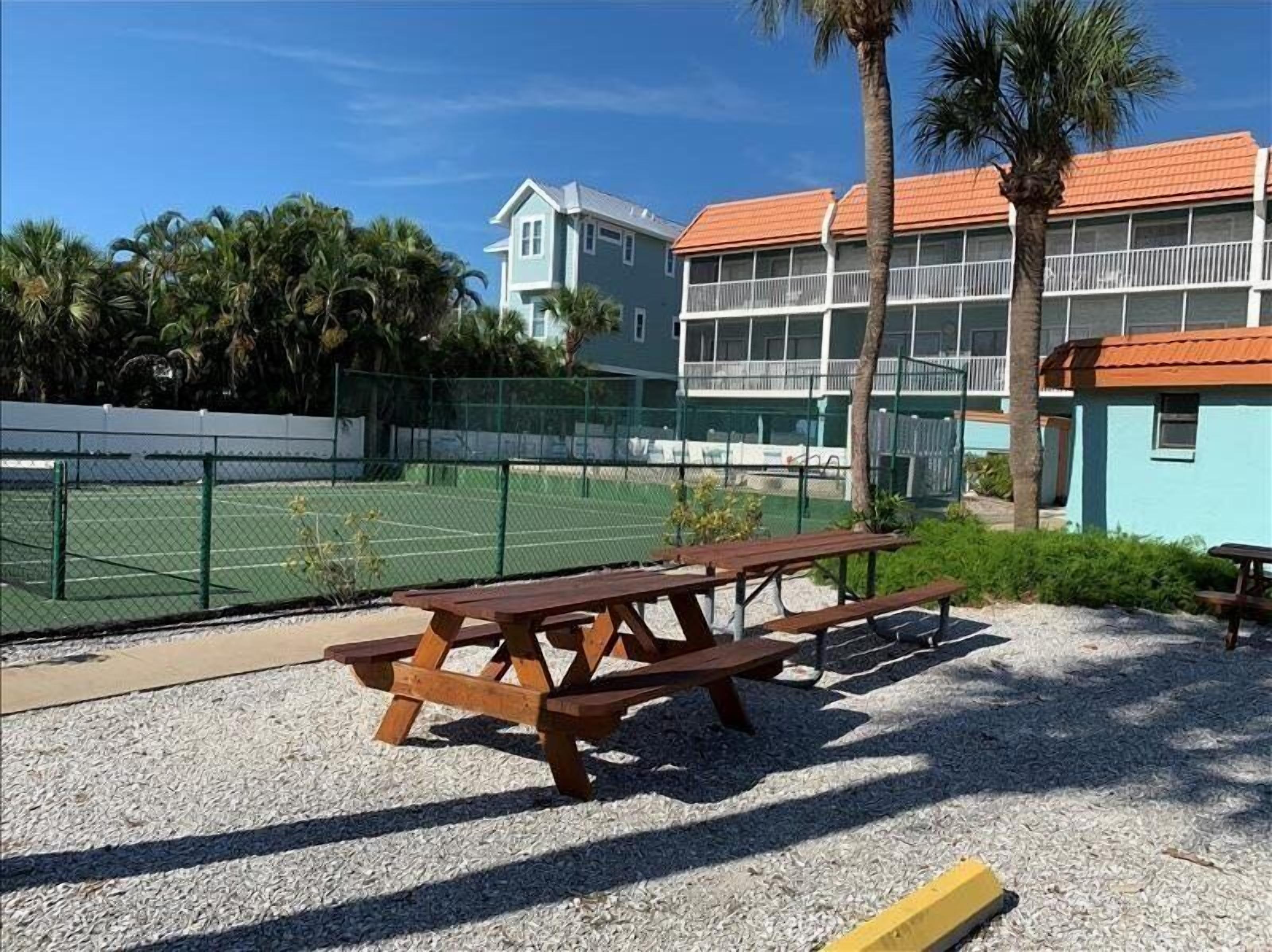 A tennis court with a picnic table and a building with a red roof in the background.