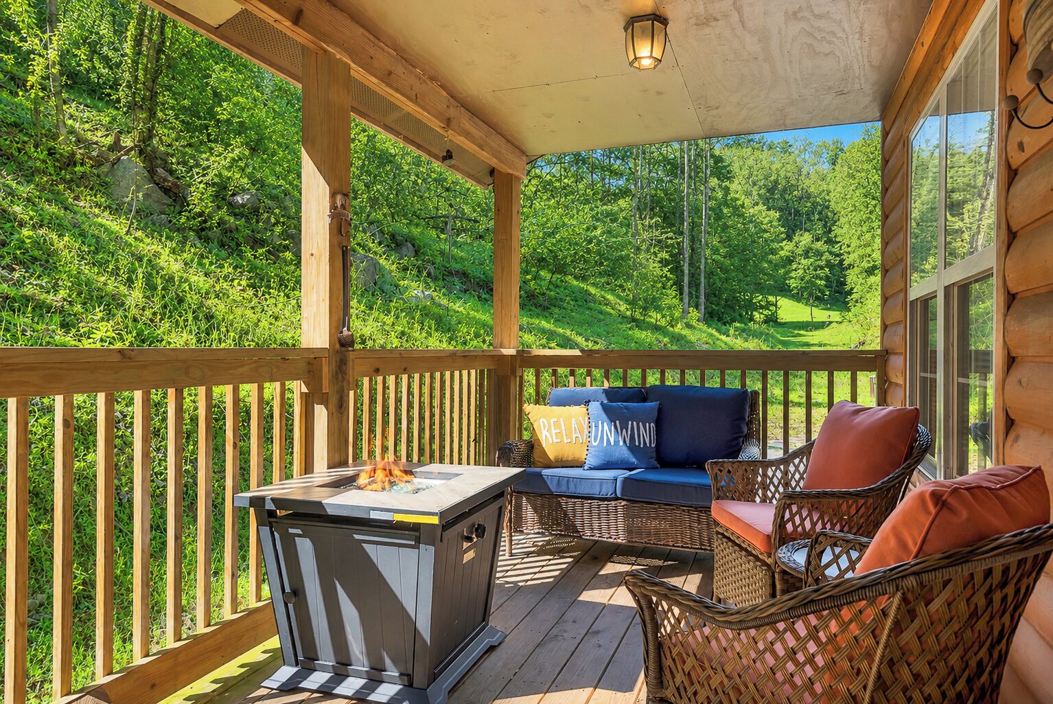 A wooden deck with a grill, wicker furniture, and a view of a forested hillside.