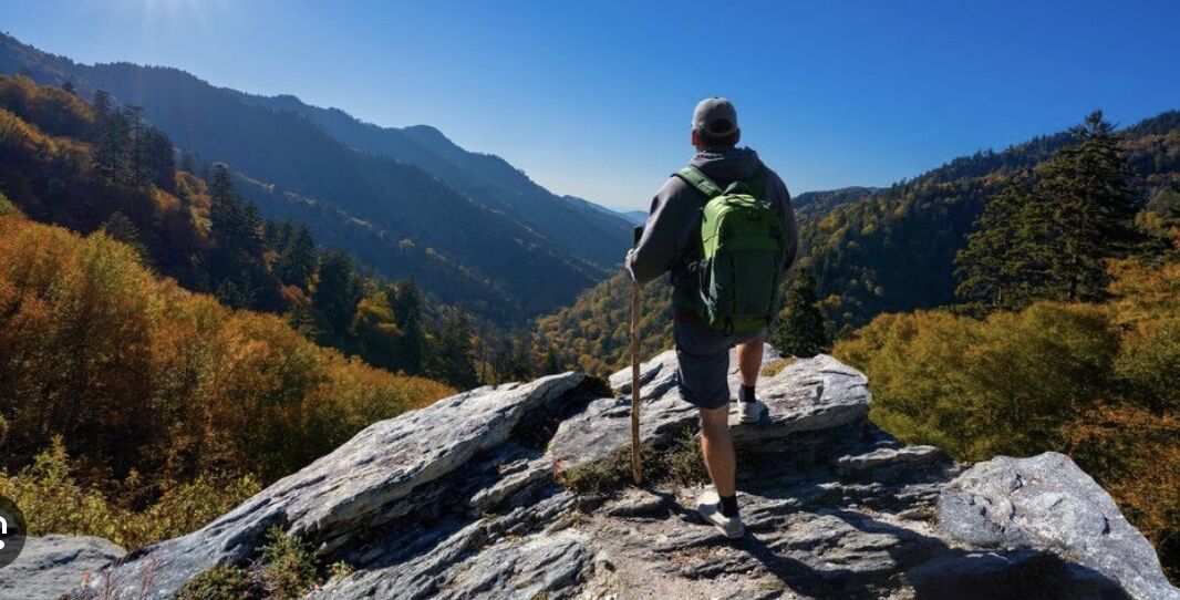 Een persoon wandelt over een rotsachtig bergpad met een rugzak en wandelstok, omringd door herfstkleuren en bergketens in de verte.
