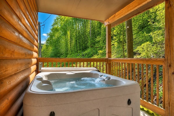 A wooden cabin with a hot tub on a deck overlooking a forest.