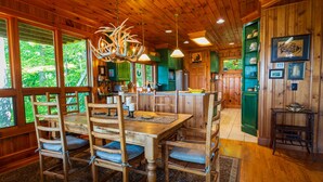 A rustic wooden interior with a dining area featuring a large wooden table and chairs, a chandelier with antlers, and a kitchen with green cabinets and a bar area. - Mountain Cabin,180* View,HotTub,SpaTub,Pool Table,WiFi (Ferguson)