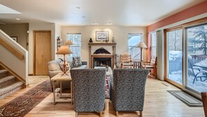 A cozy living room with a fireplace, two patterned armchairs, a coffee table, and a dining area with a view of a snowy outdoor deck. - Beautiful half duplex home, recently remodeled with garage and private hot tub!! (Frisco)